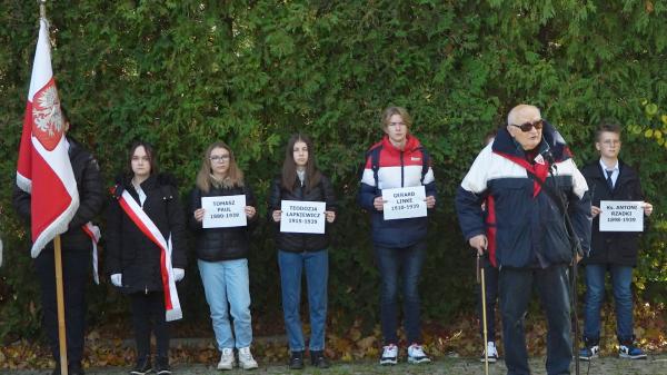 Młodzież trzymająca kartki z nazwiskami osób pomordowanych w Forcie VII stoi wzdłuż drogi prowadzącej do ściany śmierci. Przemawia członek rodziny  - kliknij aby powiększyć