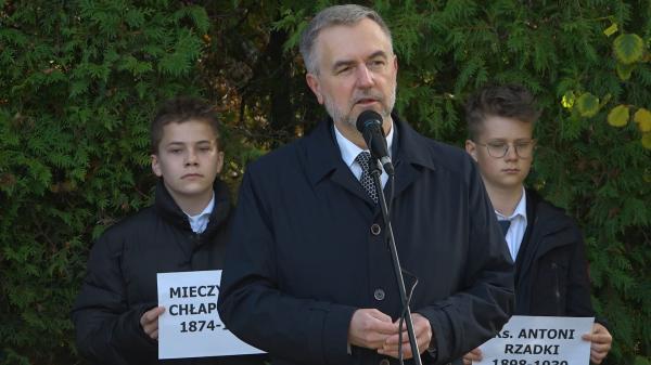Przemawia Marszałek Marek Woźniak. W tle chłopcy trzymający kartki z nazwiskami osób pomordowanych  - kliknij aby powiększyć